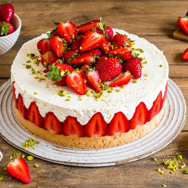 Ein Erdbeerkuchen, inspiriert von der klassischen deutschen Erdbeertorte, ist mit frischen Erdbeeren und gehackten Pistazien belegt. Geschnittene Erdbeeren säumen den Rand des Kuchens, weitere ganze Beeren und Pistazien liegen verstreut auf dem Holztisch daneben.