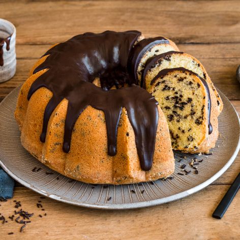 Ein Ameisenkuchen mit Schokoladenglasur liegt auf einem Teller. Er ist teilweise angeschnitten und gibt den Blick auf das helle, mit Schokoladenstückchen gesprenkelte Innere frei. Ein Becher mit Schokoladensoße, ein Messer und Schokoladenstücke stehen daneben auf einem Holztisch.
