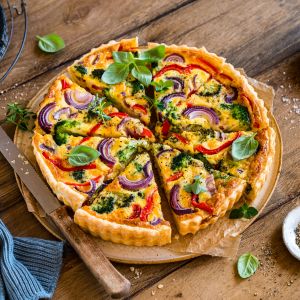 Eine bunte Gemüsequiche liegt auf Pergamentpapier auf einem Holztisch, garniert mit frischem Basilikum. Die Scheiben zeigen rote Zwiebeln, rote Paprika, Brokkoli, Kräuter und geschmolzenen Cheddar. Daneben liegen ein Messer, eine Stoffserviette und kleine Schälchen mit Salz und Pfeffer.