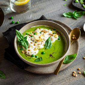Eine Schüssel grüne Erbsensuppe, garniert mit Sahne, grünen Erbsen, Pinienkernen, schwarzem Pfeffer und frischen Basilikumblättern, steht auf einem Teller mit einem goldenen Löffel. Zitronenspalten, Salz und noch mehr Basilikum stehen daneben auf einem rustikalen Holztisch.