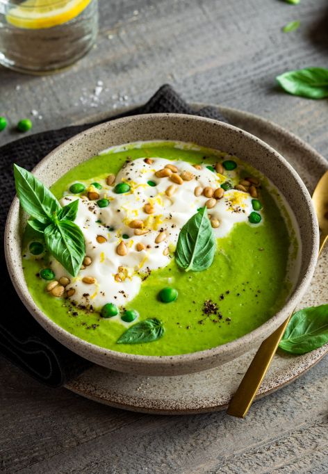 Eine Schüssel grüne Erbsensuppe, garniert mit Sahne, grünen Erbsen, Pinienkernen, schwarzem Pfeffer und frischen Basilikumblättern, steht auf einem Teller mit einem goldenen Löffel. Zitronenspalten, Salz und noch mehr Basilikum stehen daneben auf einem rustikalen Holztisch.