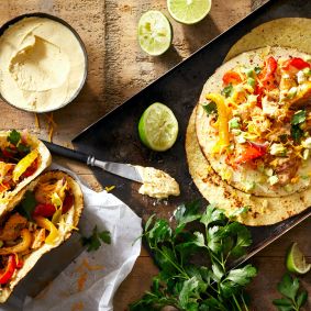 Eine Tischplatte mit Tortillas, belegt mit Hummus, gegrillten Hähnchen-Fajitas, Paprika, Käse, Petersilie und Limette. Daneben stehen eine Schüssel Hummus, frische Petersilie, Limettenhälften und ein Messer auf einer rustikalen Holzoberfläche.