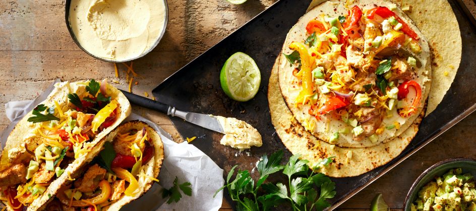 Eine Tischplatte mit Tortillas, belegt mit Hummus, gegrillten Hähnchen-Fajitas, Paprika, Käse, Petersilie und Limette. Daneben stehen eine Schüssel Hummus, frische Petersilie, Limettenhälften und ein Messer auf einer rustikalen Holzoberfläche.