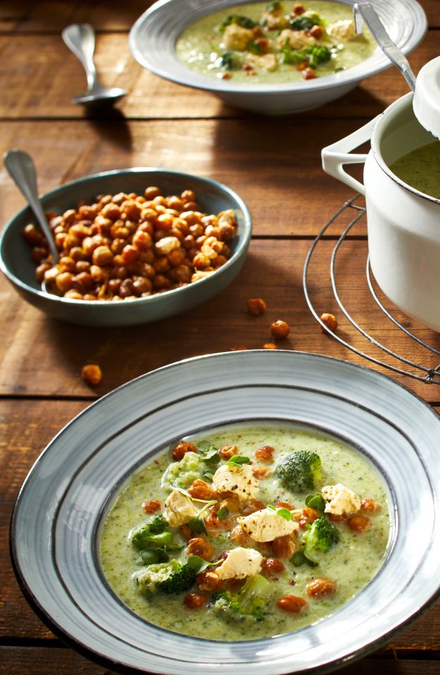 Eine Schüssel cremiger Brokkoli Suppe, garniert mit Kichererbsen und frischen Kräutern, steht auf einem Holztisch neben einem Topf Suppe, einer Schale knuspriger Kichererbsen, frischem Grün und einem Löffel.