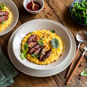 Eine Schüssel cremiges Ofenrisotto mit gelbem Cheddar, garniert mit Wurstscheiben, frischen Kräutern und rosa Pfefferkörnern auf einem Holztisch, umgeben von einer grünen Serviette, Besteck, einer Schüssel mit Gemüse, Salz und Gewürzen.