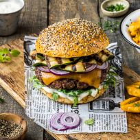 Ein Gourmet-Burger mit Cheddar-Käse, gegrillter Zucchini, roten Zwiebeln, Salat und Sauce auf einem Brötchen mit Körnern, serviert auf einem Holzbrett mit Waffelpommes, Kräutern und einer Tasse Dip-Sauce.