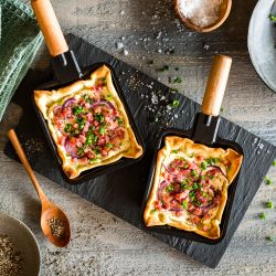 In zwei kleinen Raclettepfännchen liegt Flammkuchen: gebackenes Fladenbrot mit Sahne, Zwiebeln, Speck und Schnittlauch auf einem schwarzen Schieferbrett. Daneben liegen Salz, Pfeffer, ein Holzlöffel, noch mehr Schnittlauch und ein grünes Tuch.