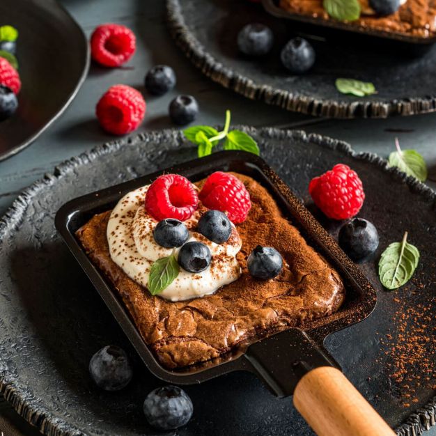 Ein quadratischer Schokoladenbrownie mit Schlagsahne, frischen Himbeeren und Blaubeeren wird in einer kleinen schwarzen Pfanne auf einem dunklen Teller serviert – ein bildschöner Leckerbissen für alle Rezeptkategorien, garniert mit zusätzlichen Beeren und Minzblättern.