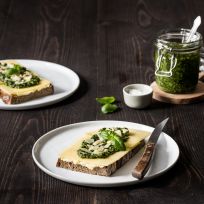 Zwei Teller mit Brotscheiben, belegt mit Kerrygold-Käse, Pesto und frischen Kräutern, stehen auf einem dunklen Holztisch. Ein Glas Pesto, ein Schälchen Salz und ein Buttermesser runden diese appetitliche Szene in der Rezeptkategorie ab.