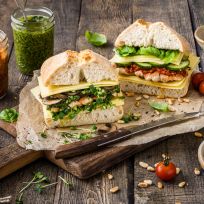 Zwei große Ciabatta schnelle Sandwiches, gefüllt mit Käse, Greens, Pilzen und gegrilltem Hähnchen, liegen auf einem Holzbrett – umgeben von Aufstrichen, Kirschtomaten, Pinienkernen und frischem Basilikum. Perfekt für jede Rezeptkategorie!.