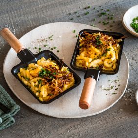 Zwei kleine Pfannen mit Käsespätzle, garniert mit knusprig gebratenen Zwiebeln und Schnittlauch, stehen auf einem runden Teller, umgeben von Schüsseln mit Salz, Pfeffer und Schnittlauch auf einem rustikalen Holztisch.