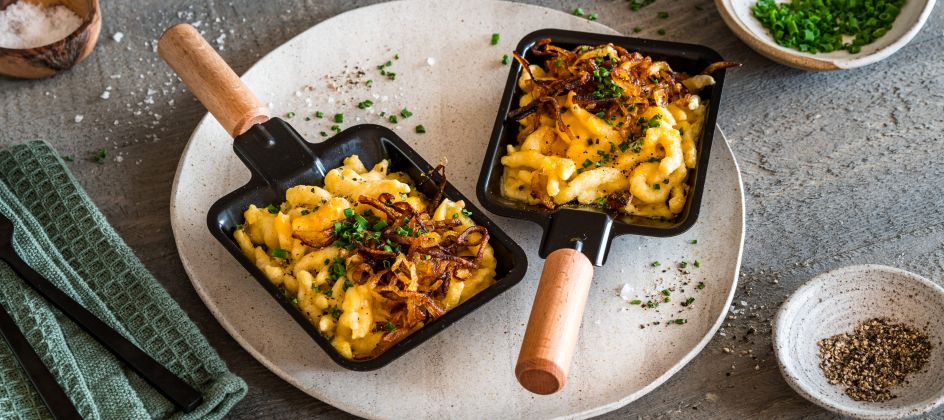 Zwei kleine Pfannen mit Käsespätzle, garniert mit knusprig gebratenen Zwiebeln und Schnittlauch, stehen auf einem runden Teller, umgeben von Schüsseln mit Salz, Pfeffer und Schnittlauch auf einem rustikalen Holztisch.