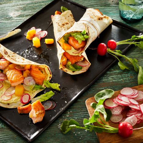 Zwei Dinkelwraps mit Lachs, belegt mit Salat, Radieschenscheiben und Mangostücken, liegen auf einem schwarzen Tablett neben einem Schneidebrett mit Radieschenscheiben, ganzen Radieschen und einem Glas Wasser auf einem rustikalen grünen Holztisch.