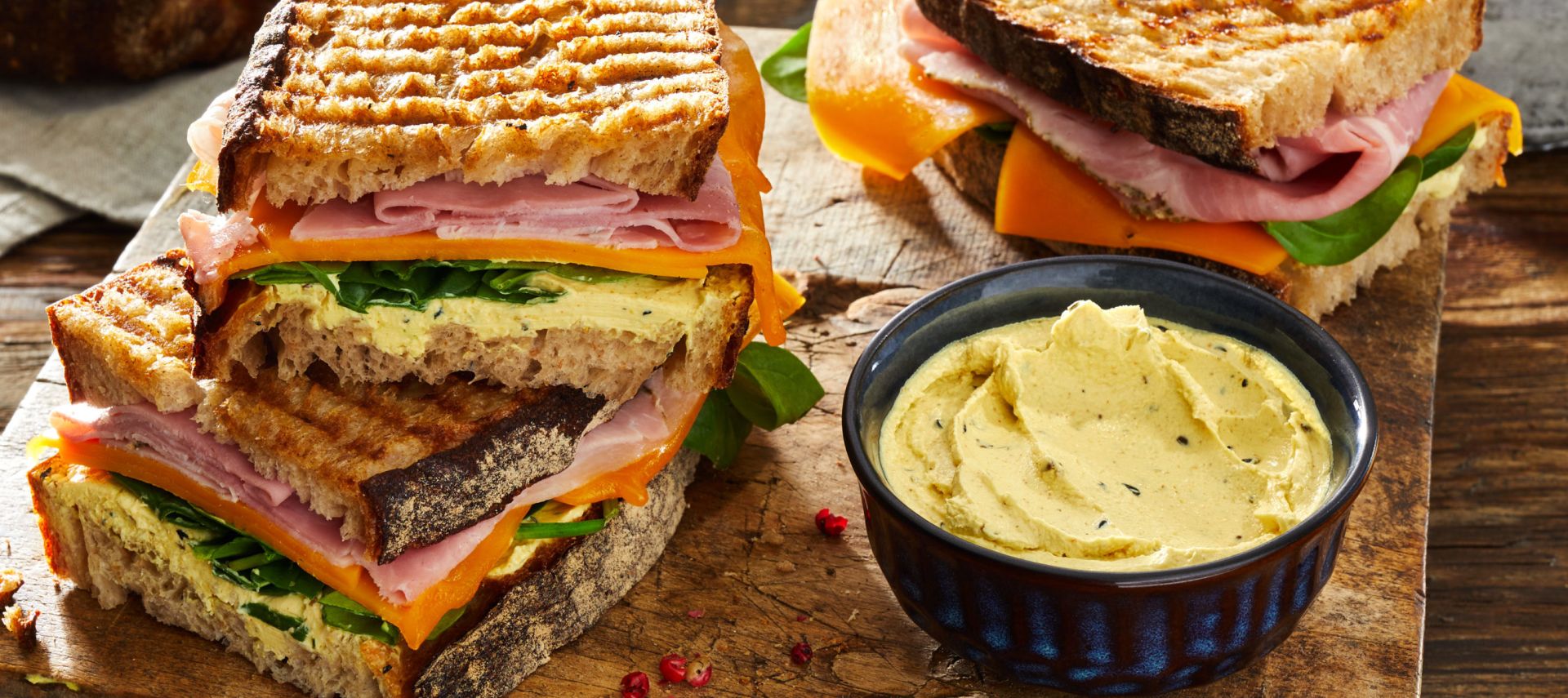 Gegrillte Schinken-Käse-Sandwiches auf einem Holzbrett gestapelt, serviert mit einer kleinen Schüssel Aufstrich, frischen Spinatblättern, rustikalem Brot und einem Kräutertopf.