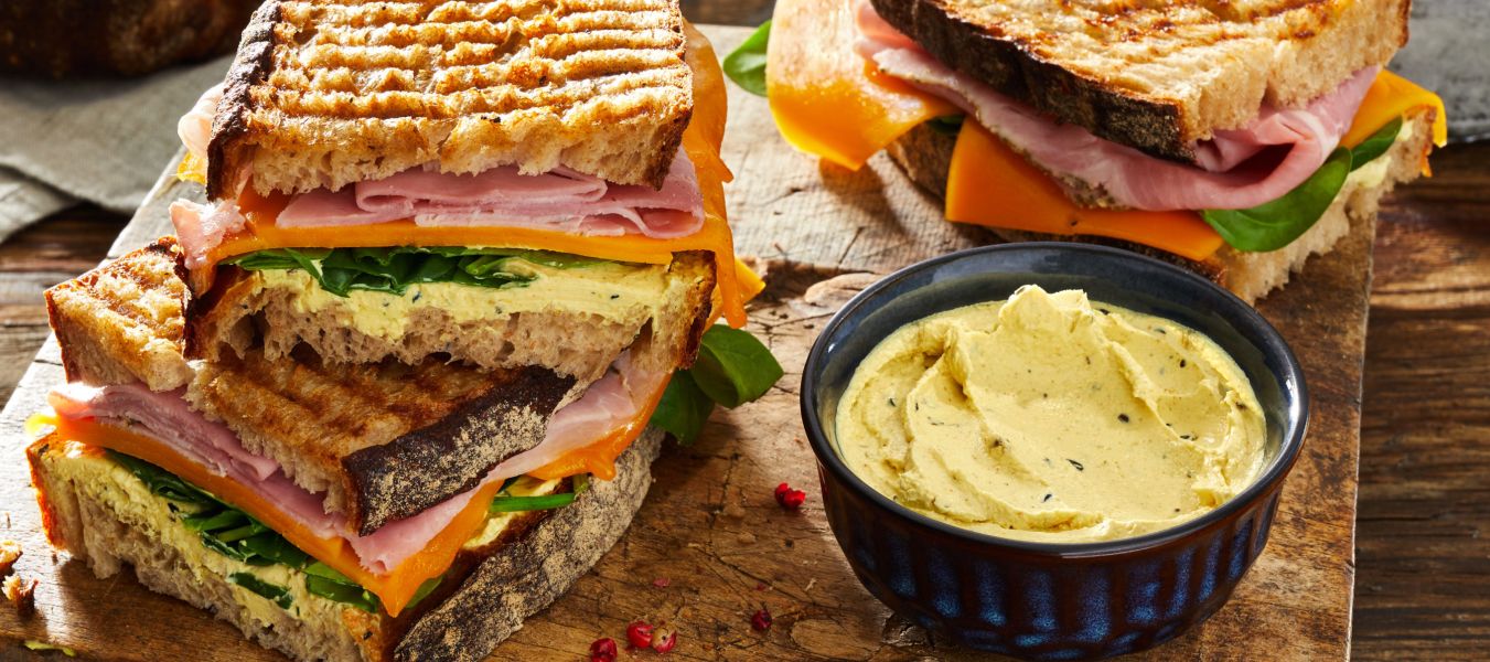 Gegrillte Schinken-Käse-Sandwiches auf einem Holzbrett gestapelt, serviert mit einer kleinen Schüssel Aufstrich, frischen Spinatblättern, rustikalem Brot und einem Kräutertopf.