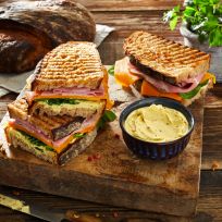 Gegrillte Schinken-Käse-Sandwiches auf einem Holzbrett gestapelt, serviert mit einer kleinen Schüssel Aufstrich, frischen Spinatblättern, rustikalem Brot und einem Kräutertopf.