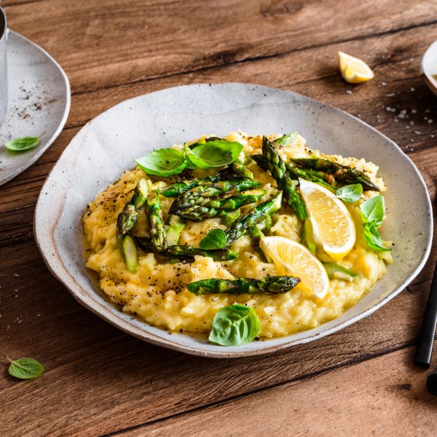 Auf einem Holztisch steht ein Teller mit cremigem Spargel-Risotto, garniert mit gegrilltem Spargel, Basilikumblättern, Zitronenspalten und schwarzem Pfeffer, dazu ein Topf Risotto, eine Gabel und ein kleiner Teller mit Zitronenscheiben.