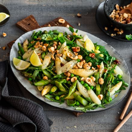 Auf einem Holztisch steht eine Platte mit warmem Spargelsalat mit Blattgemüse, Limettenspalten und gehackten Nüssen, dazu eine schwarze Serviette, Salatbesteck und eine kleine Schüssel mit zusätzlichen Nüssen.