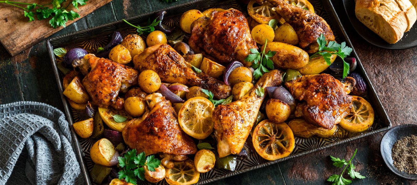 Ein Tablett mit gebratenem Hähnchen, Kartoffeln, roten Zwiebeln und Zitronenscheiben, garniert mit Petersilie und einer Prise Meersalz, steht auf einem dunklen Holztisch neben einem Teller Brot und einer kleinen Schüssel Pfeffer.