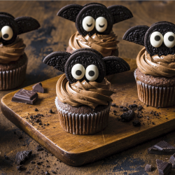 Vier Schokoladenmuffins mit Schokoladenglasur sind mit Flügeln aus Keksen und Augen aus Zucker verziert und sehen aus wie Fledermäuse. Sie werden auf einem Holzbrett mit Schokoladenstücken daneben präsentiert.
