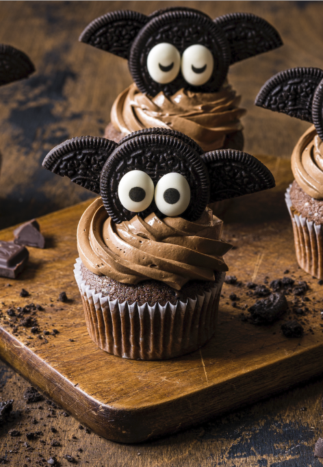 Vier Schokoladenmuffins mit Schokoladenglasur sind mit Flügeln aus Keksen und Augen aus Zucker verziert und sehen aus wie Fledermäuse. Sie werden auf einem Holzbrett mit Schokoladenstücken daneben präsentiert.