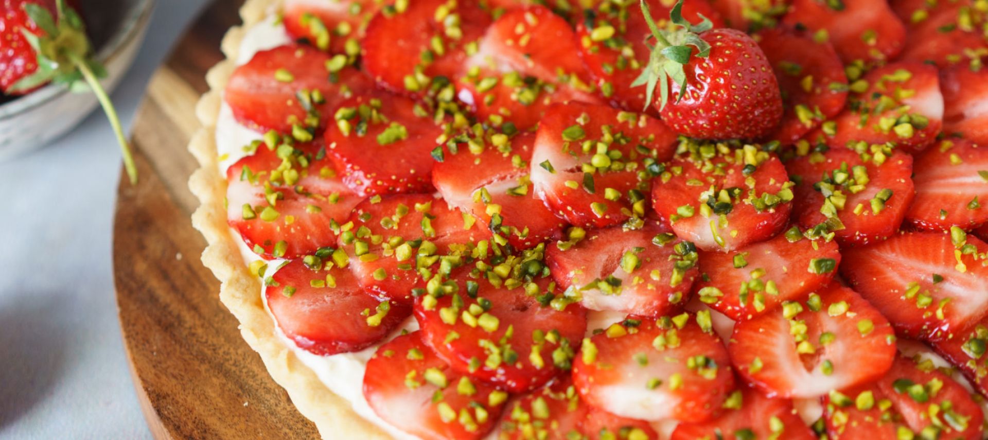Eine Erdbeertarte mit geschnittenen Erdbeeren, gehackten Pistazien und einer cremigen Mascarpone-Schicht steht auf einem Holzbrett. Daneben stehen frische Erdbeeren, ein Servierbesteck und eine kleine Schale mit Pistazien.