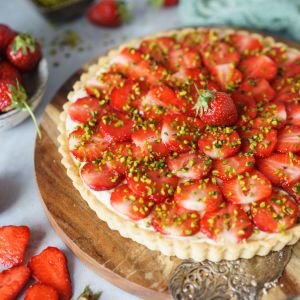 Eine Erdbeertarte mit geschnittenen Erdbeeren, gehackten Pistazien und einer cremigen Mascarpone-Schicht steht auf einem Holzbrett. Daneben stehen frische Erdbeeren, ein Servierbesteck und eine kleine Schale mit Pistazien.