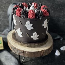 Eine Halloween-Torte aus Schokoladenkuchen, dekoriert mit Geistern aus weißem Fondant und schwarz-rotem Zuckerguss, präsentiert auf einem Holzständer. Im Hintergrund liegt eine Packung Kerrygold-Butter auf einem grauen Tuch.