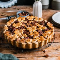 Ein Apfel-Pekannuss-Kuchen mit dekorativen Teigformen obendrauf steht auf einem Gitterrost auf einem rustikalen Holztisch, umgeben von einer Vase mit Blumen, Tellern, Gabeln und Keramiktassen.
