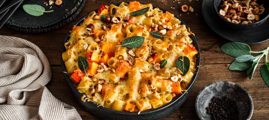 Gebackener Nudelauflauf mit Käse, geröstetem Kürbis, Salbeiblättern und gehackten Haselnüssen in einer schwarzen Schüssel auf einem rustikalen Holztisch, umgeben von einer Serviette, Tellern, Besteck und kleinen Schüsseln mit Belag.