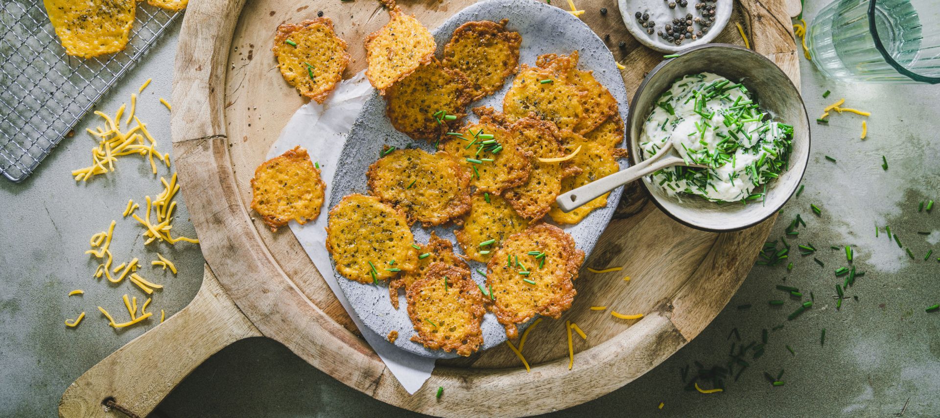 Ein Holztablett mit goldbraunen, knusprigen Krapfen auf Backpapier, garniert mit Schnittlauch. Daneben eine Schüssel cremiger Schnittlauch-Dip mit Kräutern, eine Schale schwarzer Pfeffer, Wassergläser und verstreut geriebene Cheddar-Chips.
