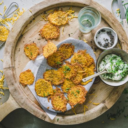 Ein Holztablett mit goldbraunen, knusprigen Krapfen auf Backpapier, garniert mit Schnittlauch. Daneben eine Schüssel cremiger Schnittlauch-Dip mit Kräutern, eine Schale schwarzer Pfeffer, Wassergläser und verstreut geriebene Cheddar-Chips.