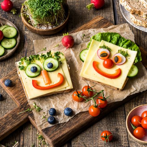 Scheiben Brot, auf denen mit verschiedenem Gemüse wie Paprika, Gurken, Tomaten und Oliven lustige Gesichter gestaltet wurden. Um den Teller herum liegt weiteres buntes Gemüse, das die Szene spielerisch und farbenfroh ergänzt.