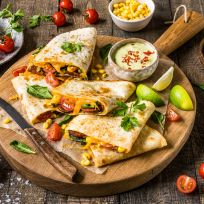 Drei Quesadilla-Ecken mit geschmolzenem Käse, Mais und Gemüse auf einem runden Holzbrett, garniert mit Koriander und Kirschtomaten, serviert mit cremiger Dip-Sauce, Limettenspalten und frischem Spinat auf einem rustikalen Holztisch.