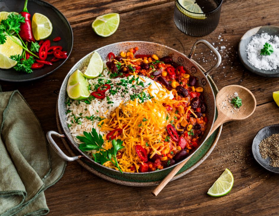 Eine Schüssel Chili sin Carne, garniert mit geriebenem Käse, Petersilie, roten Chilischeiben, serviert mit weißem Reis und Limettenspalten auf einem rustikalen Holztisch, umgeben von Gewürzschalen und einem Glas Wasser.