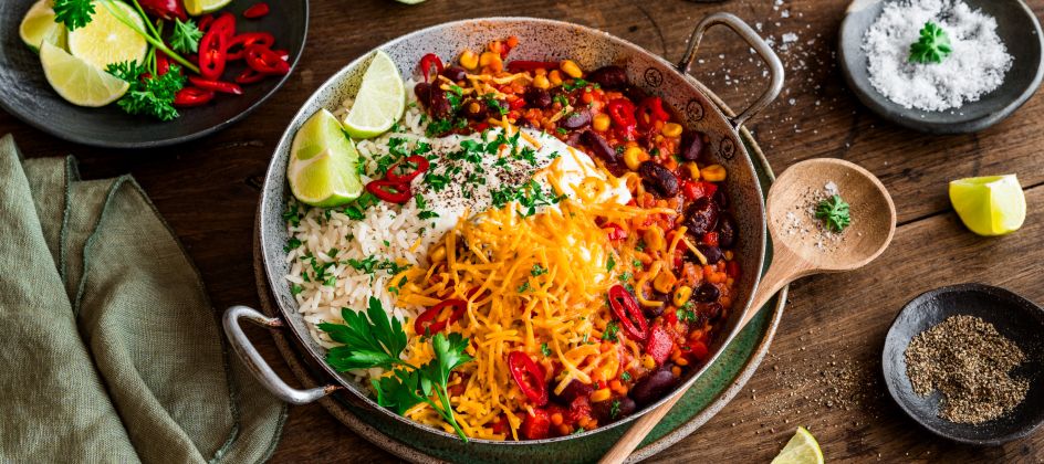 Eine Schüssel Chili sin Carne, garniert mit geriebenem Käse, Petersilie, roten Chilischeiben, serviert mit weißem Reis und Limettenspalten auf einem rustikalen Holztisch, umgeben von Gewürzschalen und einem Glas Wasser.