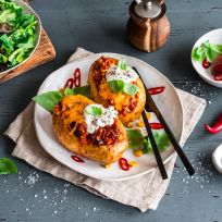 Zwei Ofenkartoffeln mit Chili con Carne, geschmolzenem Käse, Sauerrahm und Basilikum liegen auf einem Teller mit geschnittenen roten Chilischoten. Ein Beilagensalat, Salz, Pfeffer und Chiligarnitur sind auf einem rustikalen blauen Holztisch angerichtet.