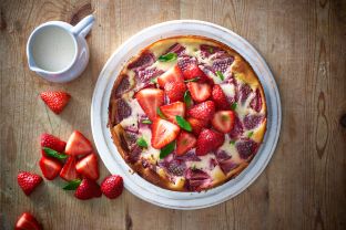 Ein köstlicher Erdbeer-Käsekuchen mit frischen Erdbeeren und Minzblättern liegt auf einem weißen Teller, mit einem Krug Sahne und verstreuten Erdbeeren auf einem Holztisch.