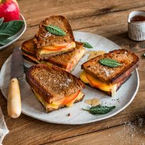 Ein Teller mit vier gegrillten Käsesandwiches und Apfelscheiben, garniert mit Salbeiblättern, steht auf einem Holztisch neben einem Messer, einer Schüssel Salz, einer Tasse Soße, frischen Äpfeln und einer beigen Serviette.