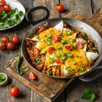 Herzhafte Enchiladas mit Hackfleischfüllung, überbacken mit Käse, serviert in einer gusseisernen Pfanne auf einem Holzbrett. Auf dem Tisch daneben liegen frische Chilis, Tomaten und Pfefferkörner, die das würzige Gericht optisch ergänzen.