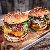 Zwei Gourmet-Chorizo-Burger mit geschmolzenem Cheddar, Rucola, Speck und geschnittenen roten Chilischoten auf Sesambrötchen, serviert auf einem mit braunem Pergamentpapier ausgelegten Metalltablett.