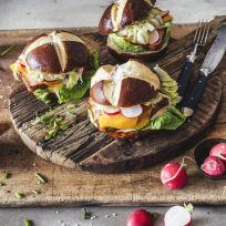 Drei Sandwiches im Biergarten-Stil mit Salat, Käse, Radieschenscheiben und Laugenbrötchen liegen auf einem rustikalen Holzbrett, umgeben von frischen Radieschen, gehacktem Schnittlauch, einer Gabel und einem Messer, einer Pfeffermühle und einem kleinen Schälchen Salz.