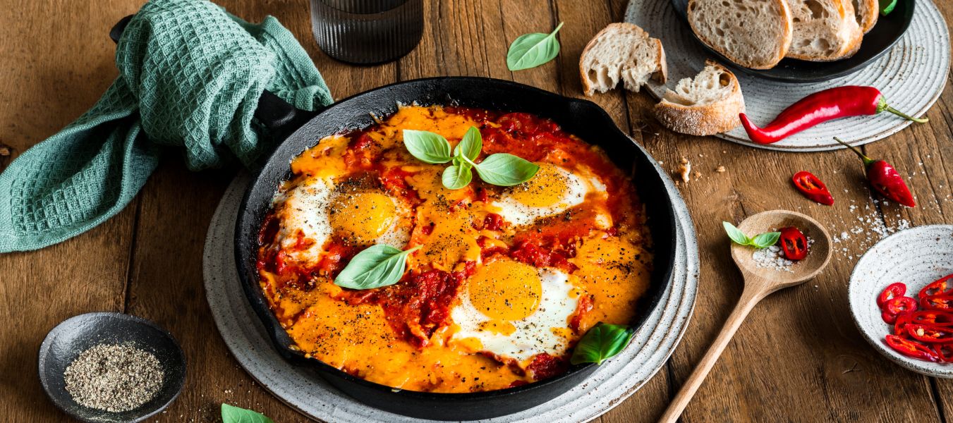 Eine Pfanne Shakshuka mit Eiern in Tomatensoße, garniert mit Basilikum und einer Prise Chili, steht auf einem Teller. Brotscheiben, ein Glas Wasser, ein Holzlöffel mit Chilischeiben und eine Schale Pfeffer stehen auf einem rustikalen Holztisch.
