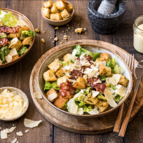 Ein Holztisch mit zwei Schüsseln Caesar-Salat, garniert mit Speck, Croutons und gehobeltem Parmesan. Daneben stehen Schüsseln mit Croutons und Käse, ein Glas Dressing, eine Pfeffermühle und Besteck.