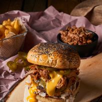 Ein BBQ-Pulled-Pork-Burger mit geschmolzenem Käse, würzigem Krautsalat und eingelegten Jalapeños auf einem Mohnbrötchen liegt auf einem Holzbrett. Daneben stehen eine Schüssel Pulled Pork und ein Korb Pommes für das perfekte Burger-Festmahl.