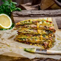 Ein Stapel Cheddar-Quesadillas mit Kräutern und saftigem Flanksteak, aus dem geschmolzener Käse quillt, liegt auf Backpapier. Im Hintergrund sind eine halbierte Zitrone, Blattgemüse und rustikale Holzscheite zu sehen, davor ein Busch.