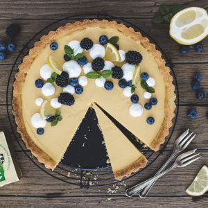 Eine Zitronentarte mit Baiser, frischen Beeren, Zitronenscheiben und Minze ruht auf einem Kuchengitter. Ein Stück fehlt, während drei Gabeln, eine Zitrone, Blaubeeren und ein Butterpäckchen auf dem rustikalen Holztisch daneben liegen.