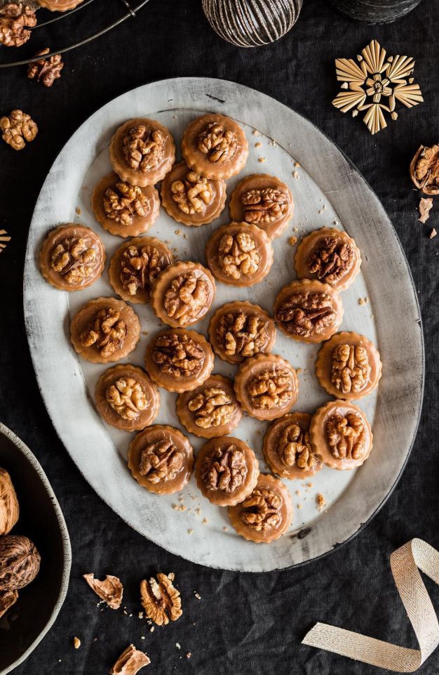 Eine ovale weiße Platte mit runden Keksen, die mit Walnusshälften belegt sind, steht auf einer dunklen Tischdecke, umgeben von ganzen Walnüssen, Walnussschalen und festlichem Schmuck – eine gemütliche Szene, inspiriert von „Kleines Kulinarium“.