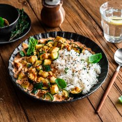 Eine schwarze Schüssel mit weißem Reis und in cremigem Frischkäse sautierten Zucchini, garniert mit frischem Basilikum und Kräutern, steht auf einem Holztisch. Daneben stehen ein Löffel, ein Glas Wasser und Gewürzgerichte.