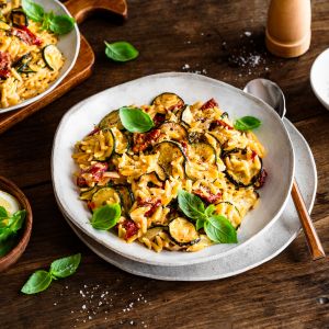 Ein Teller mit mediterranem Pastasotto mit Orzo, gerösteten Zucchini, sonnengetrockneten Tomaten und frischem Basilikum steht auf einem rustikalen Holztisch, dazu eine Schüssel mit Zitronenspalten und cremigem Frischkäse. Ein Löffel liegt neben dem Teller.